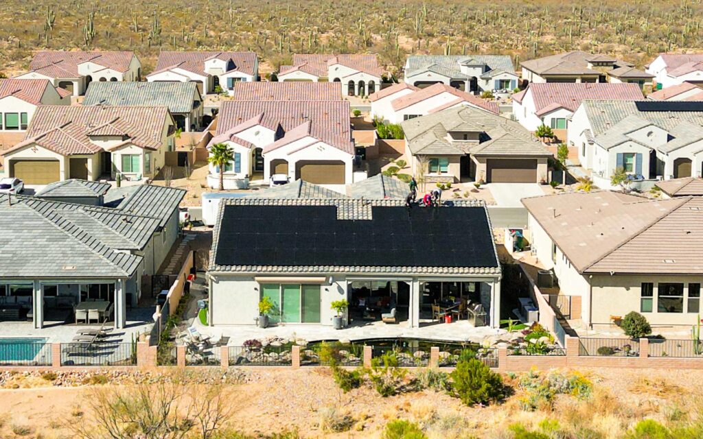 Aerial view of a suburban neighborhood with rows of houses, each with distinct roof designs and colors. The central house has a large black solar panel installation on its roof.