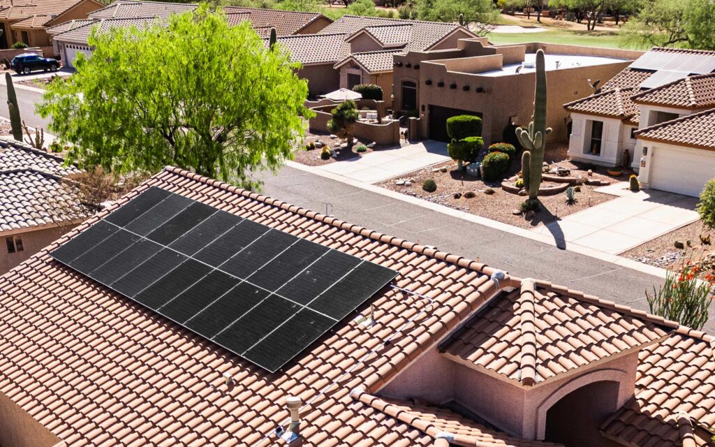 Aerial view of a residential neighborhood with a focus on a house equipped with solar panels on its roof. The house has terracotta roof tiles and is surrounded by other similar houses, desert landscaping, and cacti.