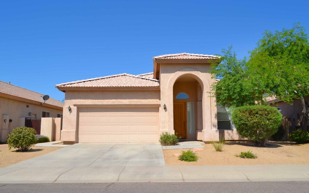 Single-story beige house with tiled roof, two-car garage, arched entryway, and desert landscaping in the front yard.