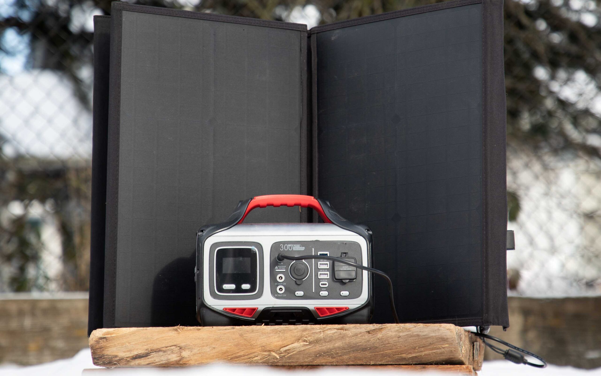 Portable solar power setup with a vertical solar panel charging a 300Watt power station outdoors, surrounded by a fence and trees.
