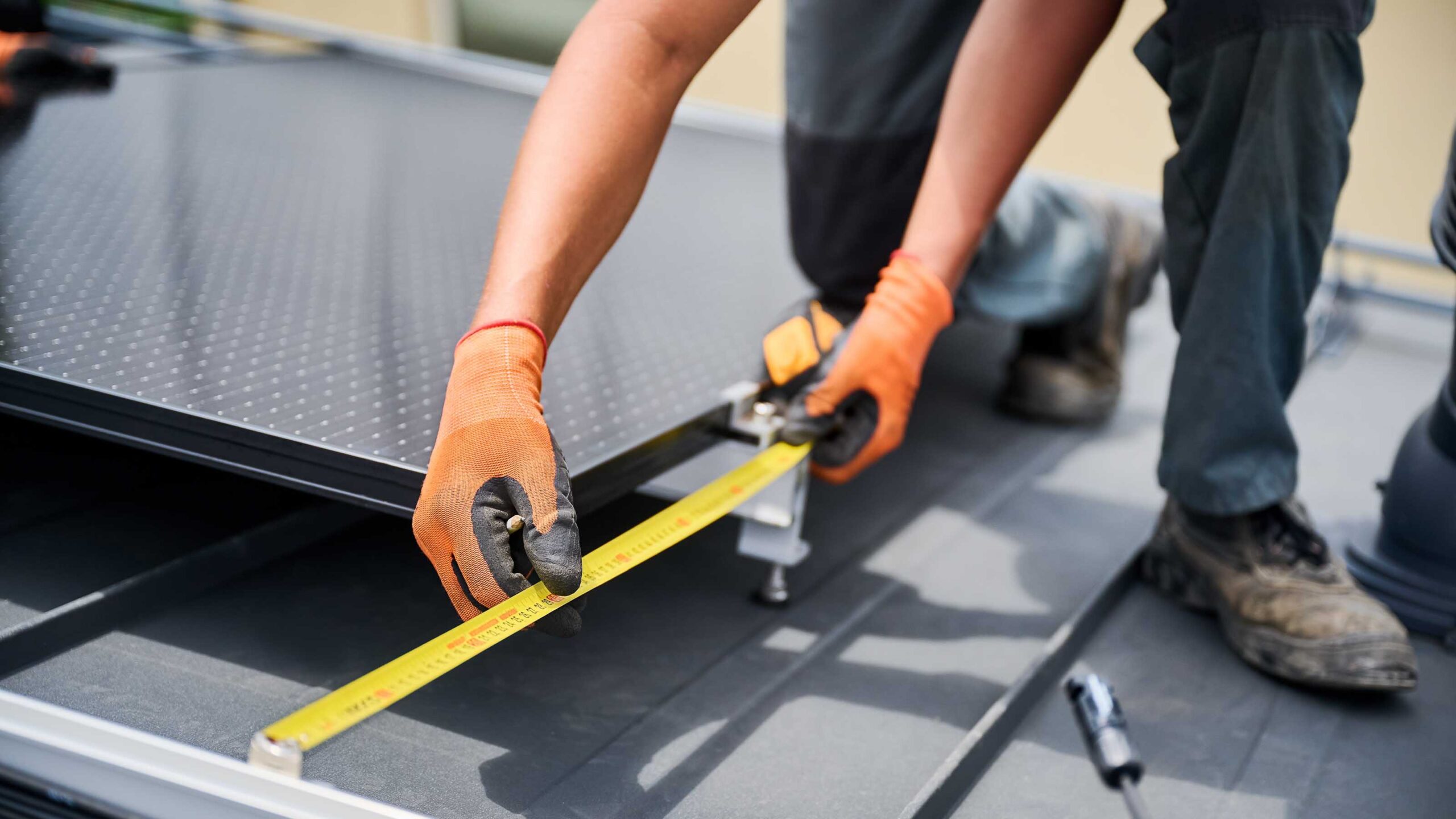 Worker wearing gloves using a tape measure to install a solar panel on a rooftop.