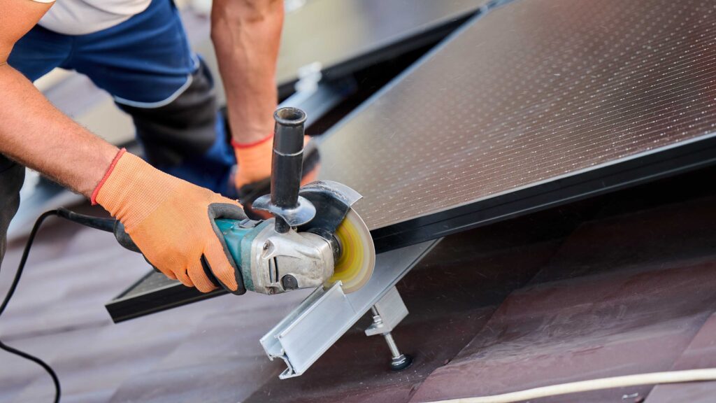 Installer wearing gloves using a power tool to secure a solar panel on a rooftop.