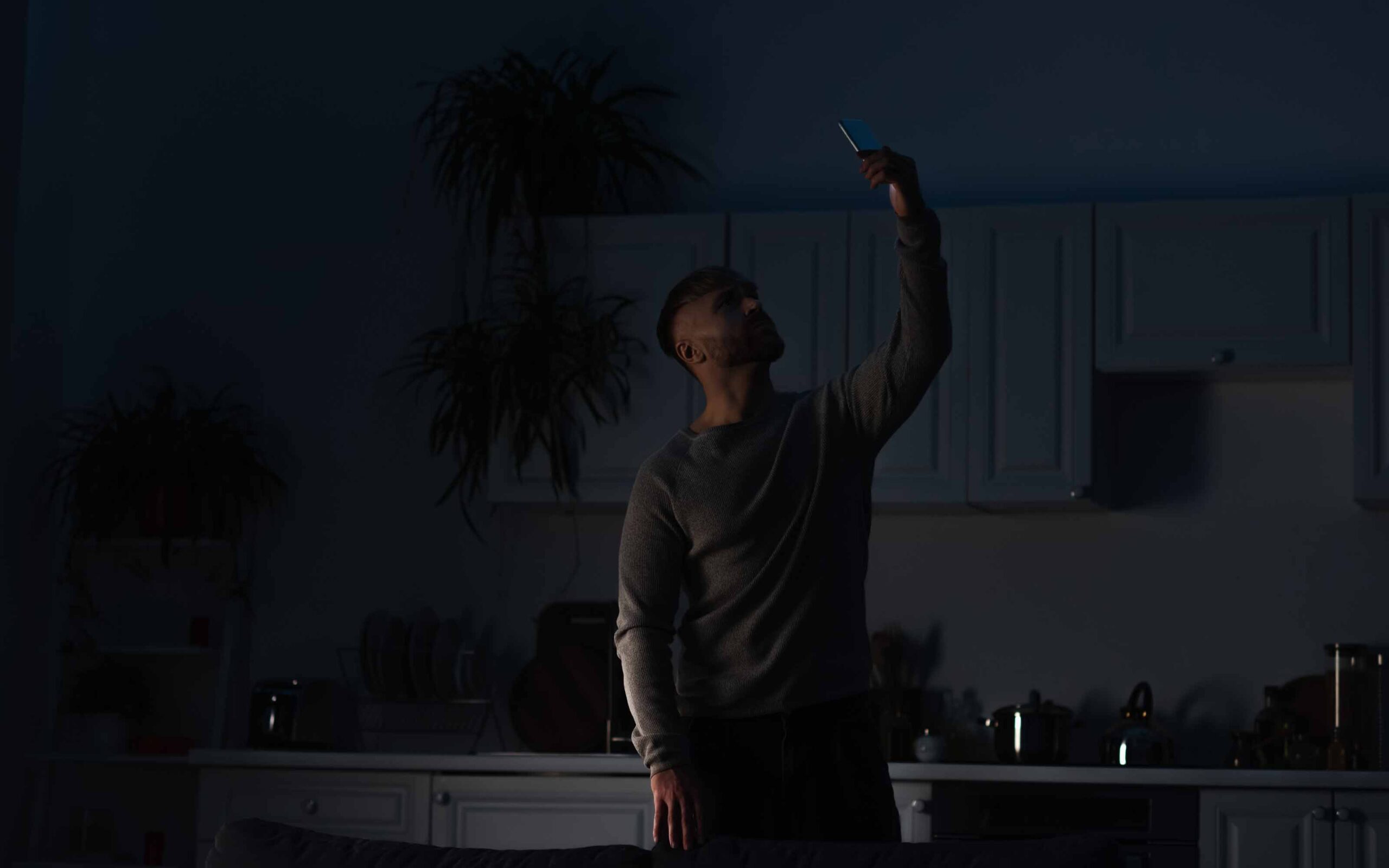 Person standing in a dimly lit kitchen, holding a smartphone aloft, surrounded by white cabinets and potted plants