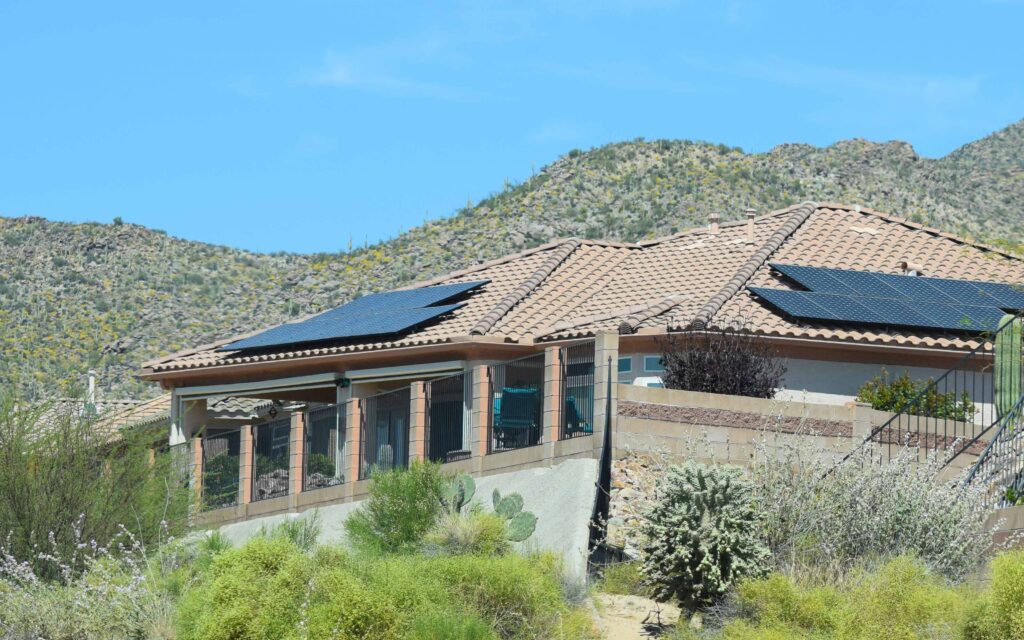 A modern desert home with solar panels installed on its terracotta tile roof, with dry, brush-covered hills in the background.